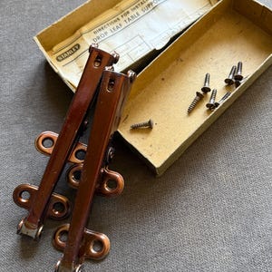May include: Two brown metal drop leaf table supports with mounting brackets and a set of small screws. The items are housed in a cardboard box labeled Stanley Directions for Installing Drop Leaf Table Support No. 1956 with instructions.
