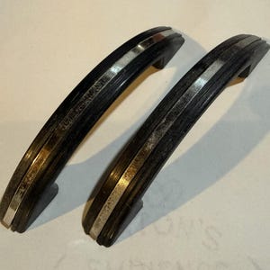 May include: Two vintage black cabinet handles with silver-colored accents. The handles have a curved, arched design with a grooved texture. The silver accents run along the center and edges of the handles, adding a touch of elegance.