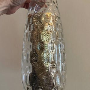 May include: A clear glass vase with a textured, dimpled exterior surface. Inside the vase, a cylindrical gold-colored metal insert features a repeating perforated pattern, creating a layered visual effect through the transparent glass walls.