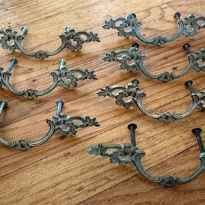 May include: A set of seven brass-toned metal drawer pulls featuring ornate, curved scrollwork designs. Each handle has two protruding mounting bolts extending from the back, with a central arched grip area and decorative wing-like ends.