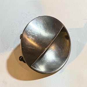 May include: A silver-toned, oval-shaped object with a diagonal divider, creating two distinct compartments. The surface has a brushed metal texture. Small, dark rivets are visible on the side. The item appears to be a vintage or antique piece.