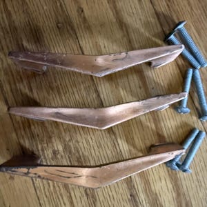 May include: Three V-shaped metal drawer pulls with a weathered copper-toned finish. Each handle features a pointed center and two mounting points. Six matching silver-colored metal machine screws are included for installation purposes.