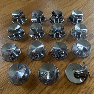 May include: A collection of fourteen circular cabinet knobs with a polished chrome finish. Each knob features a flat, reflective top surface and a cylindrical base. One knob is positioned on its side, revealing the hollow underside and mounting hole.