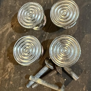 May include: A set of four circular silver-toned metal cabinet knobs, each featuring a concentric ring pattern on the face. The knobs are accompanied by four threaded metal bolts used for installation, showing a weathered and metallic finish.