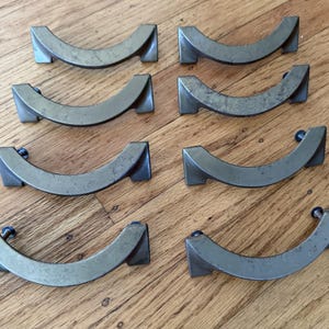 May include: Set of eight vintage metal drawer pulls. Each pull has a curved, arched design with a rectangular base on each end. The pulls are a muted, aged silver color, with small screws visible. These are likely for furniture.