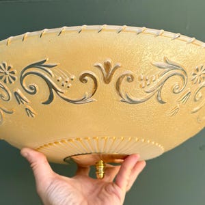 May include: A round, shallow, amber-colored glass ceiling light fixture with a textured surface. The glass features a decorative gold-toned floral and scroll pattern around the perimeter and a central gold-toned metal finial at the base.