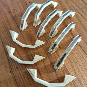May include: A collection of seven cabinet handles, each featuring an off-white, cream-colored plastic body with a central, reflective silver-toned metal inlay. The handles have an angular, geometric shape with two mounting points on each end.