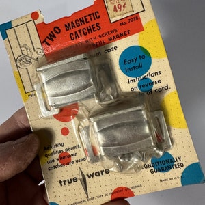 May include: Two silver magnetic catches with screws, packaged on a yellow, red, blue, and white card with the text "Two Magnetic Catches", "Complete with Screws", "Powerful Magnet", "Easy to Install", "Instructions on reverse side of card", "Conditionally Guaranteed", "True Ware", and "Made in U.S.A."