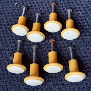 May include: Eight vintage cabinet knobs. Each knob has a yellow base, a white oval center, and a metal screw. The knobs are arranged on a dark blue textured surface.