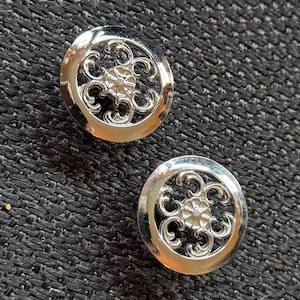 May include: Two silver-colored, round buttons with an ornate floral design. Each button features a central flower surrounded by swirling, openwork patterns. The buttons are set against a dark, textured background.