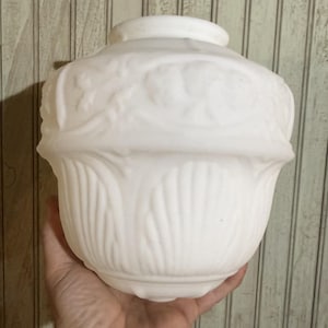 May include: A white milk glass light fixture shade with a rounded top and a flared base. The surface features a raised relief pattern with floral motifs on the upper section and vertical ribbed detailing on the lower portion of the glass.
