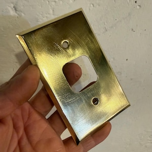 May include: A rectangular, brass-colored light switch plate with a single, square opening. The plate has two screw holes and is held in a hand against a textured, off-white wall.