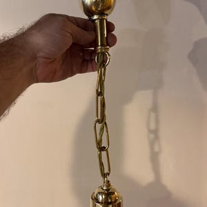 May include: A polished brass pendant light fixture featuring a bell-shaped ceiling canopy, a short chain link section, and a matching bell-shaped light socket housing. The fixture includes a braided green fabric cord threaded through the chain.