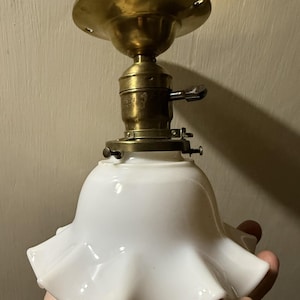 May include: A vintage-style ceiling light fixture featuring a brass mounting plate and socket with a turn-key switch. The fixture includes a white milk glass shade with a ruffled, scalloped edge, secured to the brass base by three small screws.