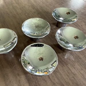 May include: Five shiny, chrome-plated cabinet knobs with a circular, concave design. Four knobs are flat, while one is elevated on a short, cylindrical base. The knobs are reflective, showing the surrounding environment.