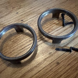 May include: Two silver metal rings with three legs each. The rings are on a wooden surface. There are four screws on the surface.