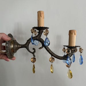 May include: An antique bronze wall sconce with two candle holders. The sconce features ornate detailing and is adorned with blue and amber-colored crystal droplets. The sconce is held by a hand.