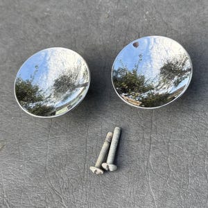 May include: Two circular cabinet knobs with a highly polished, reflective chrome finish that mirrors the surrounding environment. The set includes two threaded metal screws with a weathered, greyish appearance, positioned below the knobs.