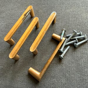 May include: Four gold-toned metal drawer pulls with a rectangular bar design and two mounting posts each. The set includes several silver-colored metal machine screws for installation. The handles show signs of wear and surface finish loss.