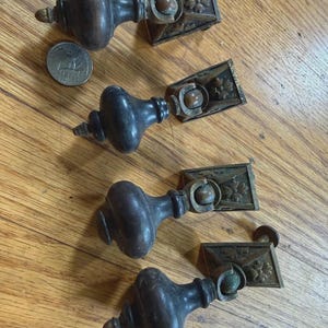 May include: Four dark brown wooden drawer pulls with brass hardware. Each pull features a rounded, bulbous handle attached to a square, floral-embossed brass backplate. A United States quarter is placed next to the top pull for scale reference.