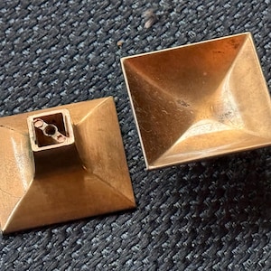 May include: Two vintage, square-shaped brass drawer pulls. Each pull has a pyramidal design with a square base and a hollowed-out interior. One pull has a square post with a screw hole.