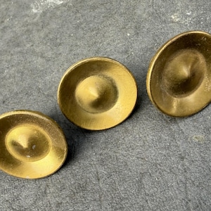 May include: Three circular brass buttons with a concave shape and a central pointed protrusion. Each button features a smooth, metallic gold-toned finish with a slightly weathered texture, arranged in a diagonal line on a dark gray surface.