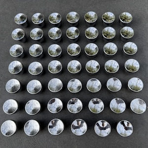 May include: A collection of forty-two small, circular, metallic buttons arranged in a grid. Each button features a highly reflective, polished chrome finish that mirrors the surrounding environment, creating a uniform and shiny appearance.