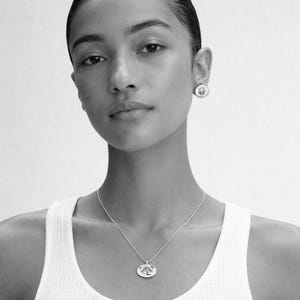 May include: A black and white image of a person wearing a silver necklace and matching earrings. The necklace features a pendant with a design. The person is wearing a white tank top.