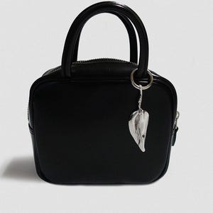 Könnte beinhalten: Eine schwarze, quadratische Handtasche mit einem oberen Griff und einem silbernen Blattanhänger. Die Tasche hat einen Reißverschluss und ein strukturiertes Design. Der Anhänger ist an einem silbernen Ring befestigt. Die Tasche ist aus glattem Material.