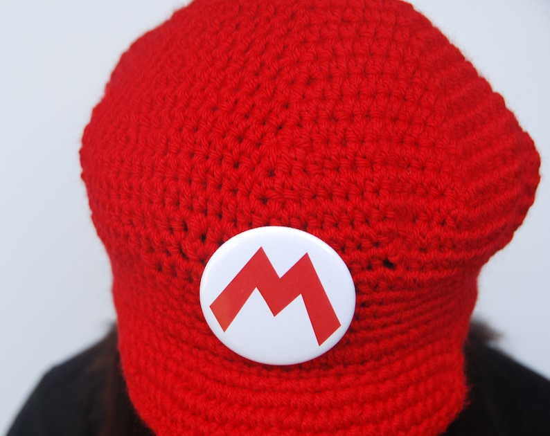 K&ouml;nnte beinhalten: Rote H&auml;kelm&uuml;tze mit einem wei&szlig;en Knopf mit einem roten 'M'-Design.