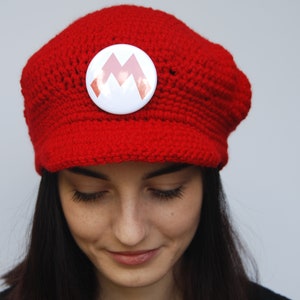 K&ouml;nnte beinhalten: Eine rote H&auml;kelm&uuml;tze mit einem wei&szlig;en Knopf, der ein rotes 'M'-Logo tr&auml;gt. Die M&uuml;tze wird von einer Person mit langen dunklen Haaren getragen.