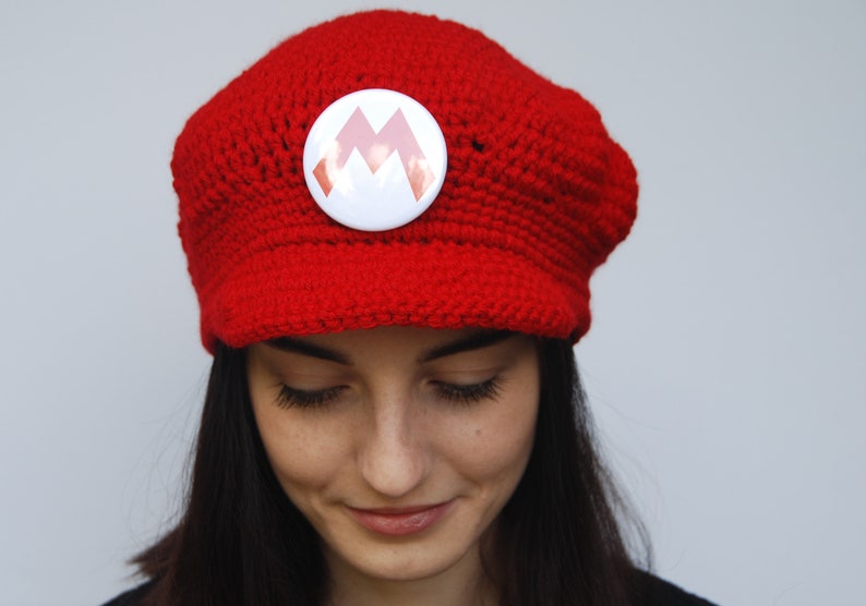 K&ouml;nnte beinhalten: Eine rote H&auml;kelm&uuml;tze mit einem wei&szlig;en Knopf, der ein rotes 'M'-Logo tr&auml;gt. Die M&uuml;tze wird von einer Person mit langen dunklen Haaren getragen.