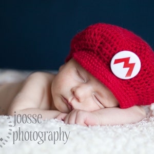 K&ouml;nnte beinhalten: Ein schlafendes Baby tr&auml;gt eine rote H&auml;kelm&uuml;tze mit einem wei&szlig;en Blitz-Emblem. Das Baby liegt auf einer wei&szlig;en Decke.