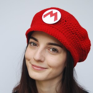 K&ouml;nnte beinhalten: Eine rote H&auml;kelm&uuml;tze mit einem wei&szlig;en M-Logo. Die M&uuml;tze hat einen Schirm und wird von einer Person mit langen dunklen Haaren getragen.