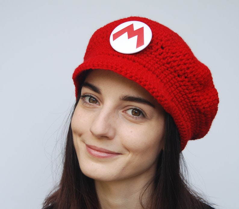 K&ouml;nnte beinhalten: Eine rote H&auml;kelm&uuml;tze mit einem wei&szlig;en M-Logo. Die M&uuml;tze hat einen Schirm und wird von einer Person mit langen dunklen Haaren getragen.