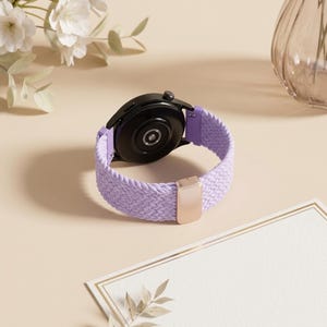 May include: A smartwatch with a black circular case and a light purple braided elastic watch band. The band features a metallic rose gold-colored rectangular sliding buckle for adjustment and light purple plastic connectors at the watch lugs.