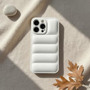 以下が含まれることがあります： 長方形のデザインの白いキルティング携帯電話ケース。ケースにはカメラの切り抜きがあり、白い縁で囲まれています。ケースは、石と乾燥した葉のあるベージュ色の表面にあります。
