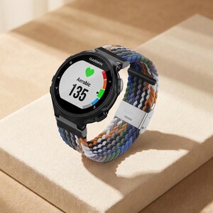 Könnte beinhalten: Eine schwarze Garmin Smartwatch mit rundem Digitaldisplay, das ein grünes Herzsymbol, den Text Aerobic und die Zahl 135 anzeigt. Die Uhr verfügt über ein mehrfarbiges, geflochtenes Stoffarmband mit silberner Metallschnalle und Verbindungsstücken.