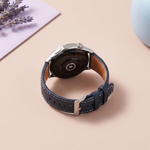 Puede incluir: Un reloj inteligente plateado con una correa de cuero azul marino y costuras blancas. La esfera del reloj es negra y la correa se asegura con una hebilla plateada. El reloj se muestra sobre una superficie rosa claro.
