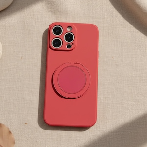 May include: A coral-coloured phone case with a circular ring holder. The case has a smooth finish and features a camera cutout with multiple lenses. The ring holder is a matching coral colour and is attached to the back of the case.