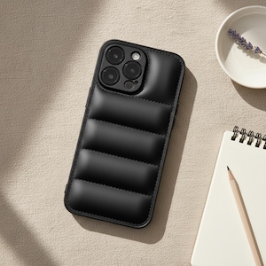 Op de afbeelding: Een zwarte gewatteerde telefoonhoes met een cameramodule. De hoes heeft een gewatteerd ontwerp met horizontale strepen. De telefoonhoes ligt op een lichtgekleurd oppervlak, met een kleine witte kom en een notitieblok met een potlood op de achtergrond.