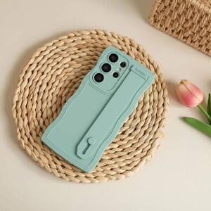 Puede incluir: Funda de teléfono de color verde azulado claro con un diseño ondulado y una correa a juego. La funda tiene múltiples recortes para la cámara y la palabra "fashion" impresa en la correa. La funda está sobre una estera tejida.