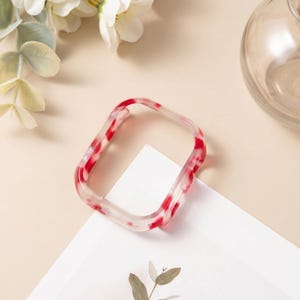 May include: A rectangular watch case cover featuring a translucent material with a marbled pattern of white and vibrant red swirls. The frame has rounded corners and a hollow center, designed to fit over the body of a digital smartwatch device.