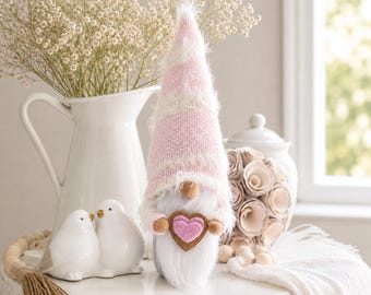 May include: A decorative gnome with a pink and white striped hat and a white beard. The gnome holds a heart-shaped ornament. The scene includes white ceramic birds, a white pitcher with flowers, and a decorative jar with paper flowers, all arranged on a white table.