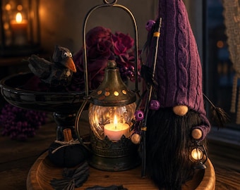 May include: A gnome with a tall purple cable-knit hat and a long black beard, holding a small lantern and a staff with purple beads. Beside it sits a metal lantern with a lit candle, a black bird figurine, and scattered black fabric pieces.