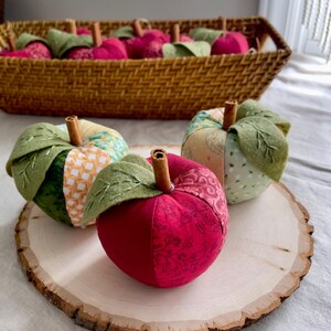 Handmade red, green and cream patchwork fabric apples displayed on a natural wood slice with a basket of red apples behind.