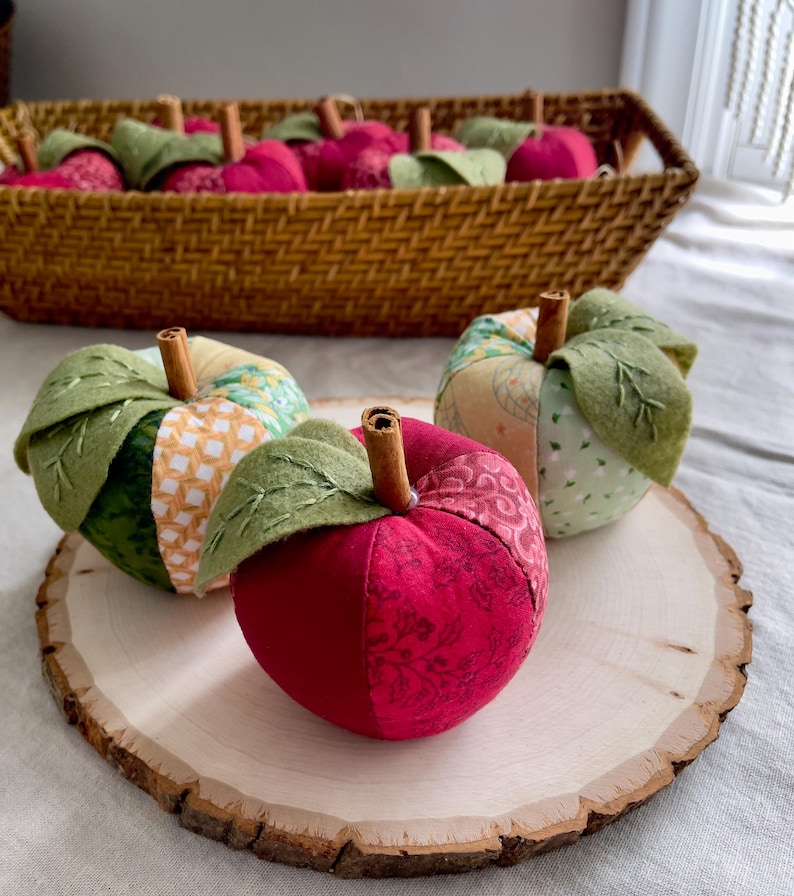 Handmade red, green and cream patchwork fabric apples displayed on a natural wood slice with a basket of red apples behind.