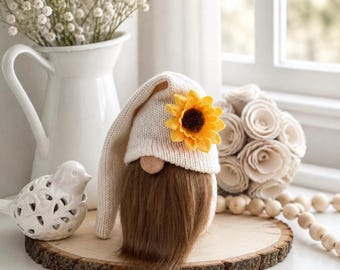 May include: A gnome figurine with a cream-colored knit hat adorned with a yellow sunflower, featuring a long brown beard and a round nose. The gnome is placed on a circular wooden slice alongside a white ceramic bird and a white flower bouquet.