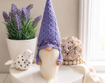 May include: A decorative gnome with a long white beard and a lavender knit hat adorned with a floral band. The gnome is on a white cake stand, with a lavender plant and a decorative bird in the background.