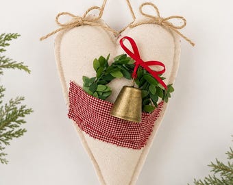 May include: A heart-shaped fabric ornament in off-white, featuring a small green wreath, a red ribbon bow, and a brass-colored bell. A strip of red and white checkered fabric is layered across the center, and a jute twine loop is attached.